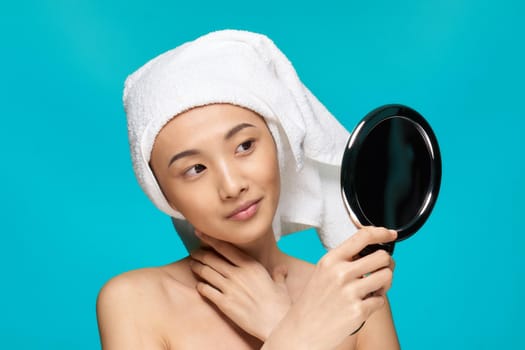 woman of asian appearance with bare shoulders looks mirror with a towel on her head blue background. High quality photo