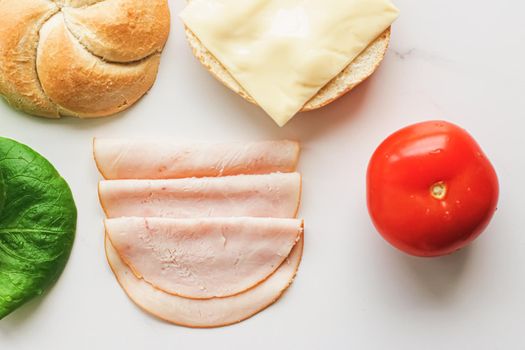 Food products and ingredients for making sandwich. Ham, cheese, burger bun, lettuce, cucumber and tomato as recipe flatlay on marble kitchen table background