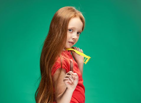 girl with red hair holding yellow glasses in her hands green background studio red t-shirt. High quality photo