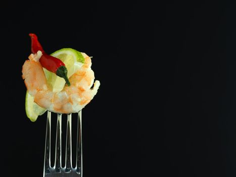 Prawn peeled boiled on a fork with a slice of lime and red chilli, isolated on a black background. High quality photo