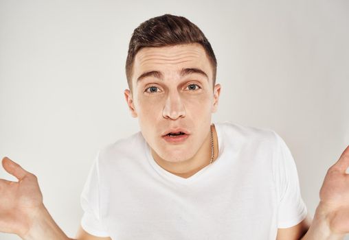 A man in bewilderment gestures with his hands on a light background in a white T-shirt cropped view. High quality photo