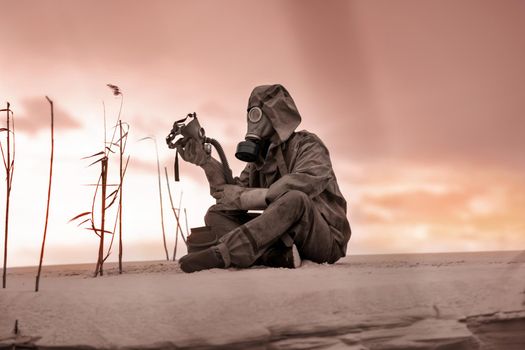 A man in a gas mask and protective suit sits on the sand and holds the gas mask of a dead friend in his hand. A man in a desperate situation. Complete loneliness. Radiation and chemical pollution.