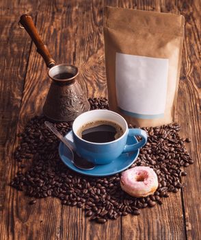 blue cup of coffee, doughnuts, beans, turkish coffee pot, and craft paper pouch bag on wooden background