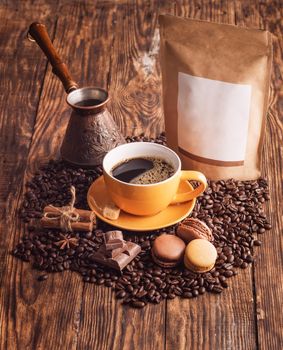 yellow cup of coffee, macaroons, beans, turkish coffee pot, and craft paper pouch bag on wooden backgroun