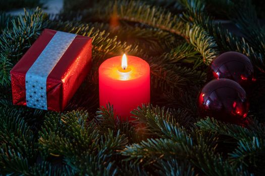 Atmospheric christmas motif with red burning candle surrounded by Nordmann fir branches, red christmas tree balls and gift parcel