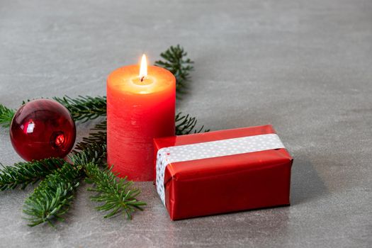 Christmas motif with red burning candle,  Nordmann fir branches, gift parcel and red christmas tree balls on a dark grey marbled  background with free space for text