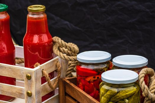 Wooden crate with glass jars with pickled red bell peppers and pickled cucumbers (pickles) isolated. Jars with variety of pickled vegetables. Preserved food concept in a rustic composition.