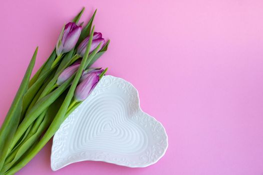 Delicate lilac tulips and a white heart-shaped plate on a pink background. Greeting card, wallpaper, background. Happy Mother's Day, Easter, Valentine's Day, or wedding.