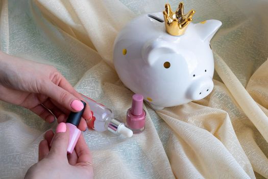 White piggy bank-piggy and female hands with pink nail design. Pink shiny nail polish manicure. Women's hands on a background of beige tulle.