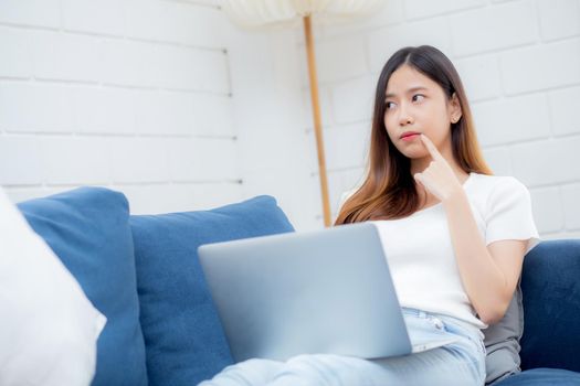 Beautiful young asian woman working on laptop computer and thinking idea on sofa at home, freelance girl sitting on couch using notebook to internet at living room, one person, lifestyle concept.