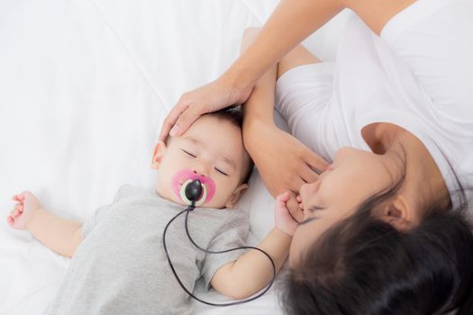 Young asian mother touch head of little baby girl with tender on bed in the bedroom, mom love newborn and care, woman with expression with child together, parent and daughter, family concept.