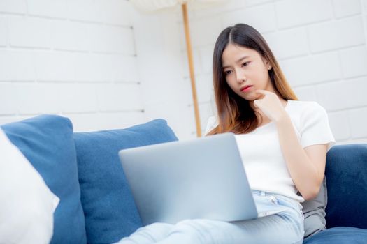 Beautiful young asian woman working on laptop computer and thinking idea on sofa at home, freelance girl sitting on couch using notebook to internet at living room, one person, lifestyle concept.