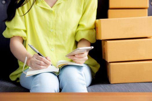 Closeup young asian woman is entrepreneur looking phone and writing order of customer with online on notebook, online shopping, logistic and delivery, female is seller working on sofa, business SME.