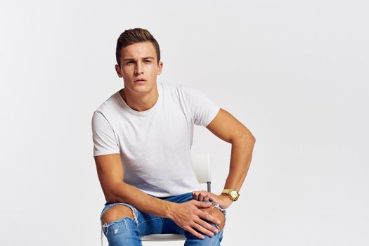 Man on a chair indoors torn jeans white t-shirt handsome face model light background. High quality photo