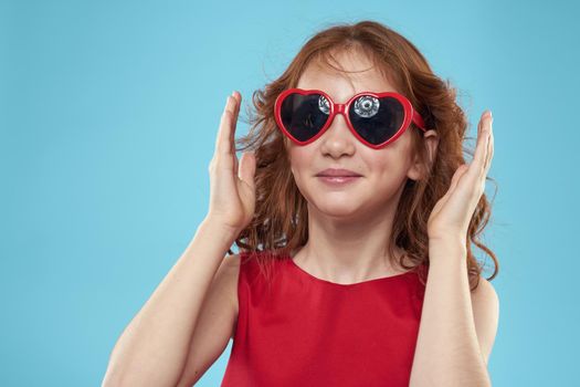 Little girl in sunglasses in the form of hearts red dress curly hair blue background. High quality photo