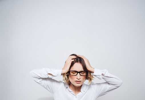 A woman in glasses and in a white shirt touches her head with her hands on a light background. High quality photo