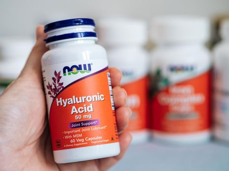 Moscow, Russia - March 09, 2021: Jar with Hyaluronic Acid capsules in female hand and others nutrition supplements in blurred background. White and orange plastic jar Hyaluronic Acid by Now Foods.