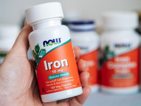Moscow, Russia - March 09, 2021: Jar with Iron capsules in female hand and others nutrition supplements in blurred background. White and orange plastic jar Iron 18mg by Now Foods.