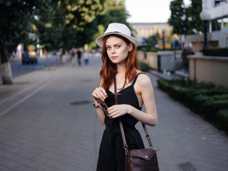 A pretty woman in a dress and hat with a bag on her shoulder walks down the street. High quality photo
