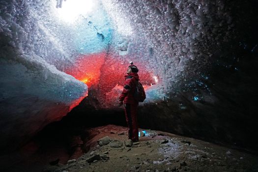 A guy in an ice cave with a lantern light. The caver descended into the ice cave. Snow stalactites and ice walls. In some places there are stones. Colored lantern beams. Bogdanovich Glacier, Almaty