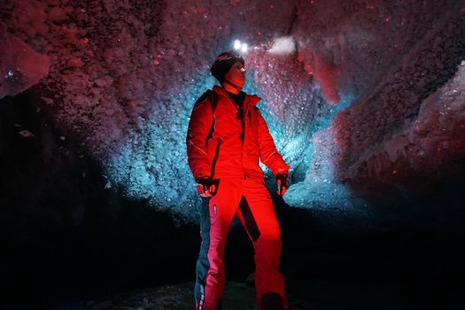 A guy in an ice cave with a lantern light. The caver descended into the ice cave. Snow stalactites and ice walls. In some places there are stones. Colored lantern beams. Bogdanovich Glacier, Almaty