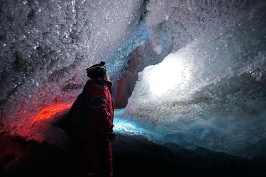 A guy in an ice cave with a lantern light. The caver descended into the ice cave. Snow stalactites and ice walls. In some places there are stones. Colored lantern beams. Bogdanovich Glacier, Almaty