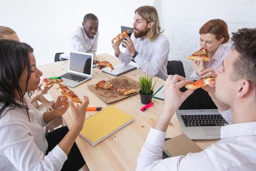 Group of diverse business people eating pizza at break while working together in an office sitting around desk