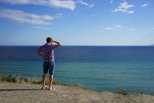 a tall, blond man standing on the edge of the cliff and looks into the distance on blue sea