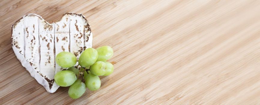 Love cheese concept, Neufchatel cheese shaped like heart with grapes on wooden cutting board , top view flat lay with copy space for text