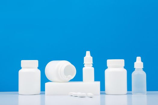 White plastic bottles with different medications, pills, nose spray and eye drops on white table against blue background with copy space. Concept of health care and pharmacy