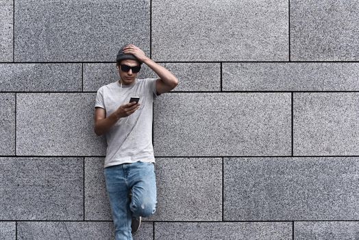 People, technology, travel and tourism - Hipster man wearing hat, smartphone on city street and listening to music over gray wall background