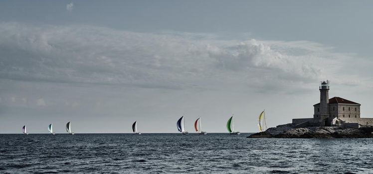 Sailboats compete in a sail regatta at sunset, Boats bend around the island with a beacon, a race, multicolored spinnakers, number of boat is on aft boats, island is on background