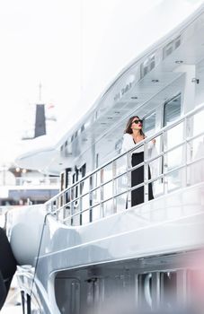 A glamorous diva in an evening dress of black color and sunglasses stands on the top deck of a huge yacht in anticipation, port Hercules, Monaco