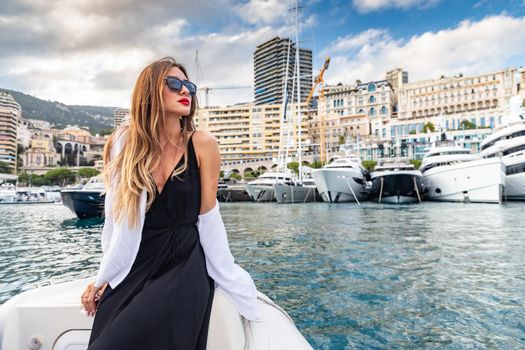 The elegant girl dressed in an evening dress of black color and sunglasses on the boat carry to the big yacht, gorgeous woman, sexual red lips, Decollete, Monaco, Monte-Carlo, is evening