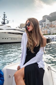The elegant girl dressed in an evening dress of black color and sunglasses on the boat carry to the big yacht, gorgeous woman, sexual red lips, Decollete, Monaco, Monte-Carlo, is evening