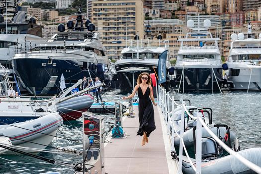 Monaco, Monte-Carlo, 27 September 2019: The elegant girl dressed in an evening dress of black color and sunglasses runs on pier in Monaco Yacht Show, gorgeous woman, sexual red lips, people