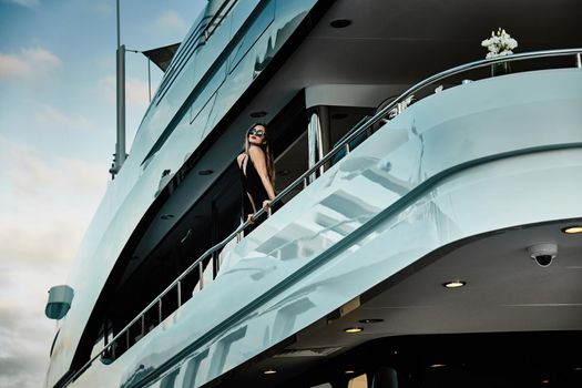 A glamorous diva in an evening dress of black color and sunglasses stands on the top deck of a huge yacht in anticipation, port Hercule, Monaco