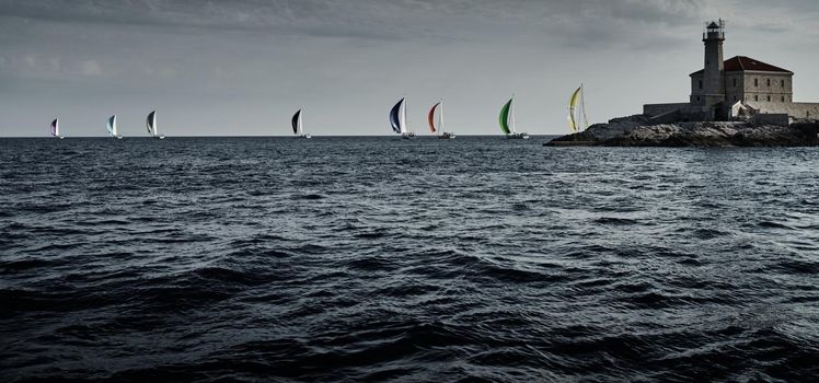 Sailboats compete in a sail regatta at sunset, Boats bend around the island with a beacon, a race, multicolored spinnakers, number of boat is on aft boats, island is on background