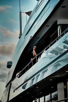 A glamorous diva in an evening dress of black color and sunglasses stands on the top deck of a huge yacht in anticipation, port Hercule, Monaco