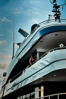 A glamorous diva in an evening dress of black color and sunglasses stands on the top deck of a huge yacht in anticipation, port Hercule, Monaco