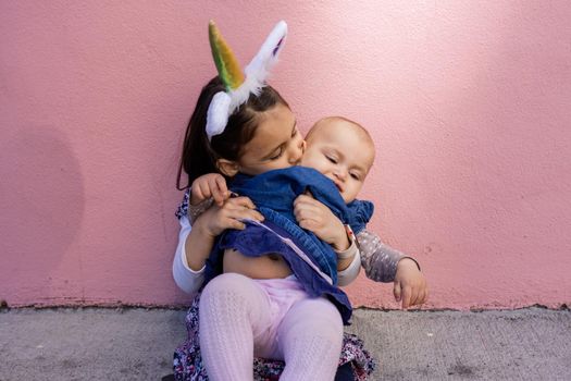 Adorable view of little girl wearing unicorn headband hugging and kissing her baby sister with pink background. Portrait of cute children sitting in front bright pink wall. Lovely kids in costumes