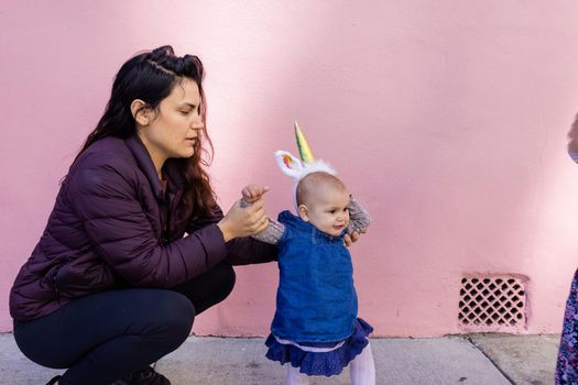 Portrait of happy mother in squatting position holding baby with unicorn headband in front of pink wall. Lovely view of smiling woman next to cute baby girl in blue dress. Happy family outdoors