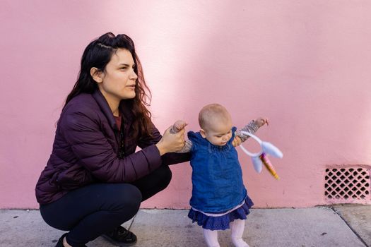 Portrait of happy mother in squatting position holding baby with unicorn headband in front of pink wall. Lovely view of smiling woman next to cute baby girl in blue dress. Happy family outdoors