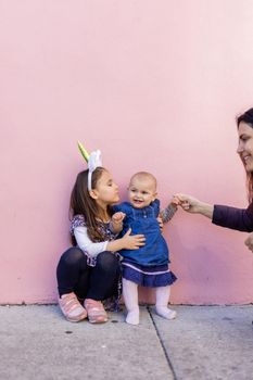 Adorable view of little girl wearing unicorn headband hugging her happy baby sister with pink background. Mother holding cute toddler hand while sitting in front bright pink wall. Lovely kids outdoors