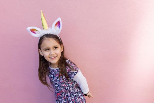 Adorable view of happy little girl wearing unicorn headband with pink background. Portrait of cute smiling child with unicorn horn and ears in front bright pink wall. Lovely kids in costumes