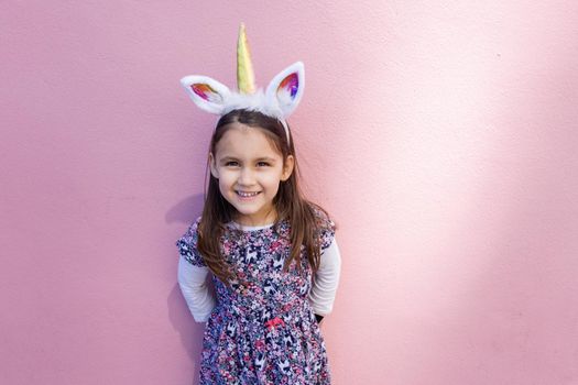 Adorable view of happy little girl wearing unicorn headband with pink background. Portrait of cute smiling child with unicorn horn and ears in front bright pink wall. Lovely kids in costumes