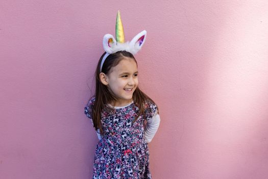 Adorable view of happy little girl wearing unicorn headband with pink background. Portrait of cute smiling child with unicorn horn and ears in front bright pink wall. Lovely kids in costumes