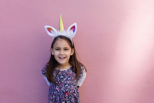 Adorable view of happy little girl wearing unicorn headband with pink background. Portrait of cute smiling child with unicorn horn and ears in front bright pink wall. Lovely kids in costumes