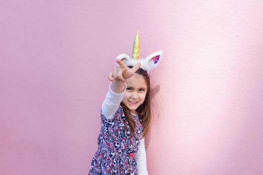 Adorable view of happy little girl wearing unicorn headband with pink background. Portrait of cute smiling child with unicorn horn and ears pointing at the camera. Lovely kids in costumes