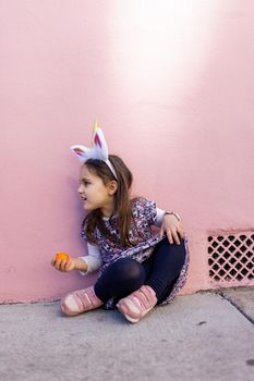 Adorable view of happy little girl wearing unicorn headband with pink wall as background. Portrait of cute smiling child with unicorn horn and ears sitting on the sidewalk. Lovely kids in costumes